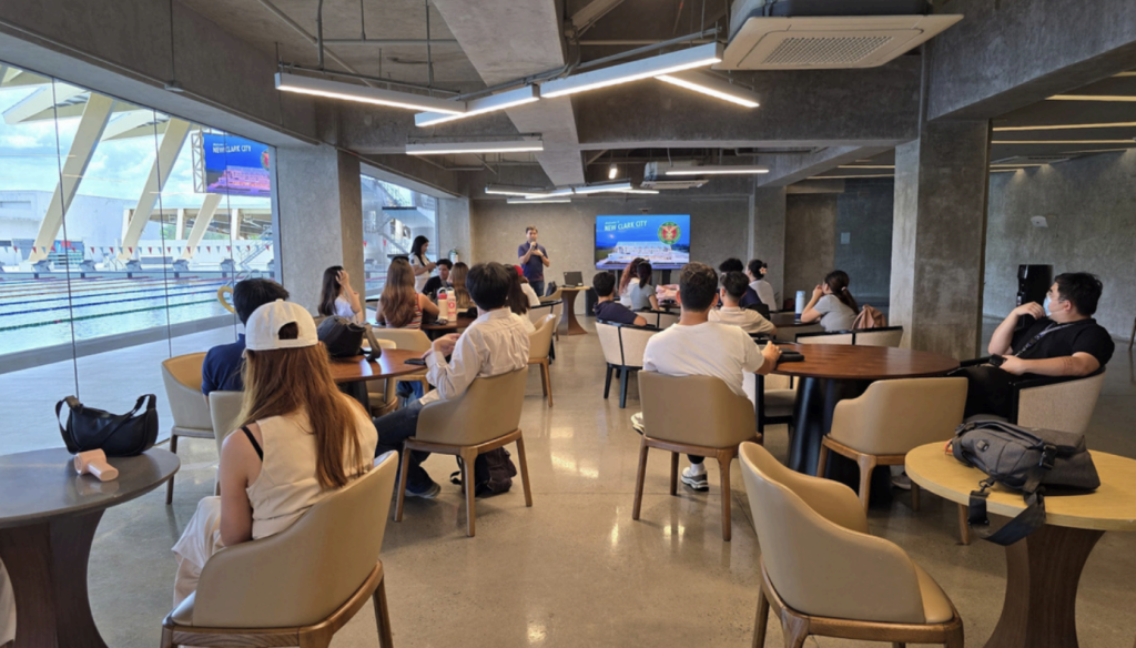 Inside an enclosed area beside the pool at the New Clark City Aquatics Center, students of PA 152 MWV are seated in a relaxed manner after a series of outdoor stopovers. A BCDA officer stands near a television screen, conducting a short briefing with visual aids. The students appear calm and attentive, some sipping water or resting, as sunlight filters in through wide glass windows.